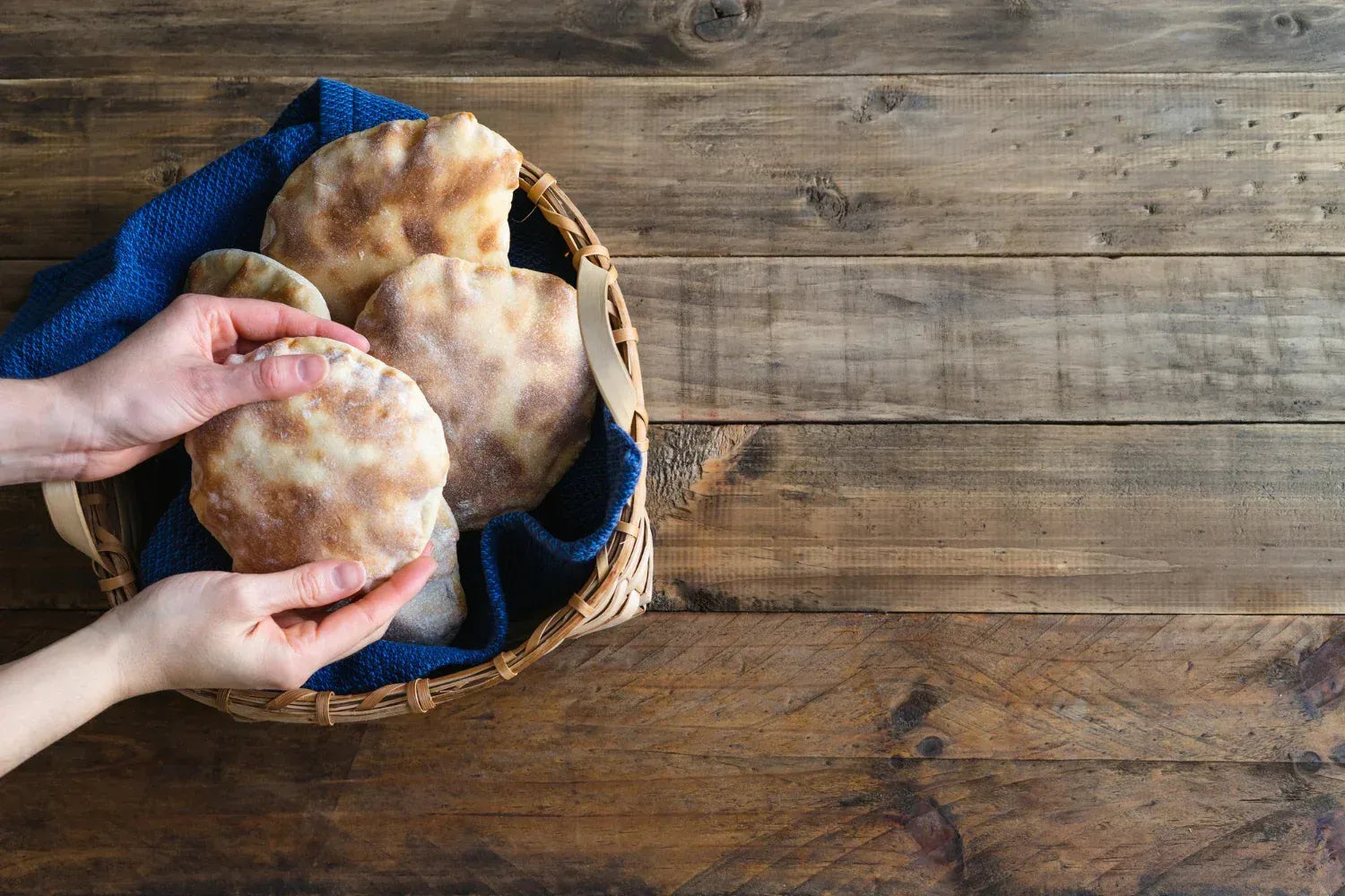 Hands holding a pita bread over a basket with more bread on a wooden surface